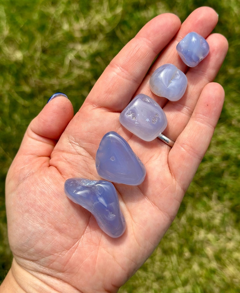 May include: A collection of six smooth, polished blue chalcedony stones of varying shapes and sizes. The stones are held in an open hand against a backdrop of green grass. The stones have a translucent quality.