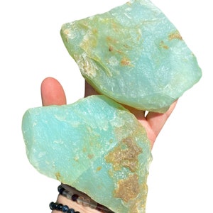 Puede incluir: Dos piedras grandes, en bruto, de color aguamarina con parches marrones se sostienen en una mano. Las piedras tienen una forma natural e irregular. Varias pulseras de cuentas son visibles en la mu&ntilde;eca debajo.