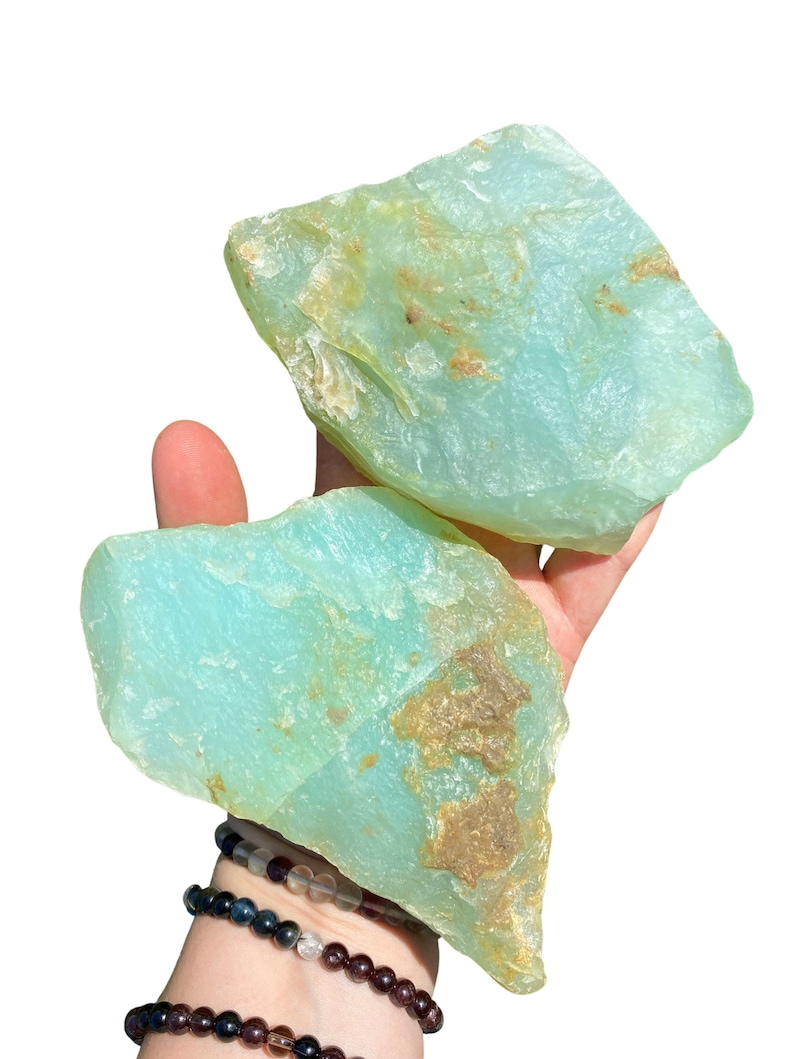 Puede incluir: Dos piedras grandes, en bruto, de color aguamarina con parches marrones se sostienen en una mano. Las piedras tienen una forma natural e irregular. Varias pulseras de cuentas son visibles en la mu&ntilde;eca debajo.