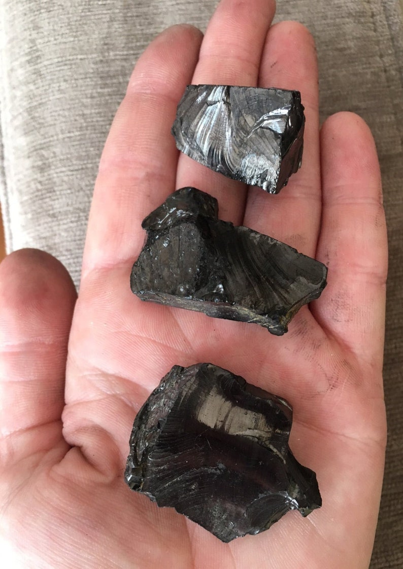 Pu&ograve; includere: Tre pezzi di roccia di ossidiana nera e lucida sono tenuti in una mano. L'ossidiana ha una superficie vetrosa e riflettente e sembra essere rotta in forme irregolari. Le rocce sono di un colore nero intenso.