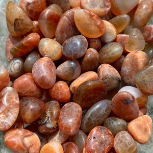 May include: A collection of polished carnelian stones in shades of orange, brown, and tan. The tumbled gemstones vary in size and shape, with smooth, rounded surfaces. The stones are displayed in a clear bowl, showcasing their natural colors and patterns.