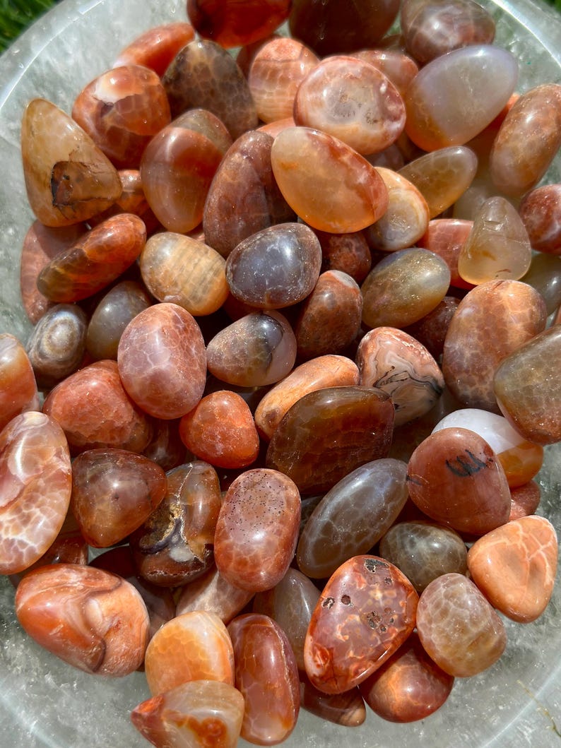 May include: A collection of polished carnelian stones in shades of orange, brown, and tan. The tumbled gemstones vary in size and shape, with smooth, rounded surfaces. The stones are displayed in a clear bowl, showcasing their natural colors and patterns.