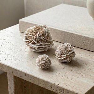 May include: Three beige desert rose stone formations of varying sizes are displayed on a light-colored stone surface. The formations have a layered, textured appearance, resembling a cluster of petals. A neutral-toned book is in the background.
