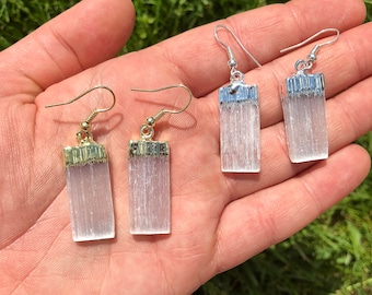 Puede incluir: Un par de pendientes de cristal de selenita con detalles dorados y plateados. Los pendientes son de forma rectangular y tienen un aspecto blanco y translúcido.