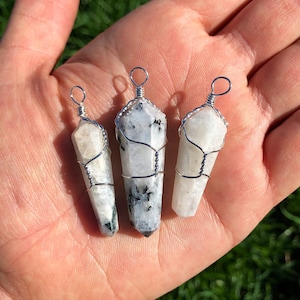 May include: Three white rainbow moonstone crystal pendants wrapped in silver wire. The pendants are different sizes and are displayed on a hand.