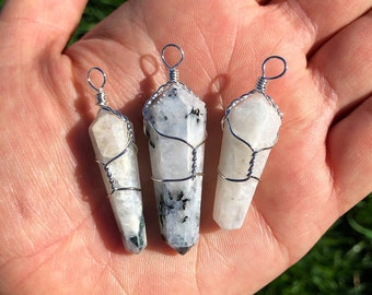 May include: Three white rainbow moonstone crystal pendants wrapped in silver wire. The pendants are different sizes and are displayed on a hand.