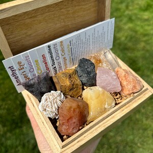 May include: A wooden box filled with various crystals and stones, including amethyst, tiger's eye, clear quartz, citrine, carnelian, black tourmaline, orange calcite, and yellow calcite. The box is open and the crystals are visible.