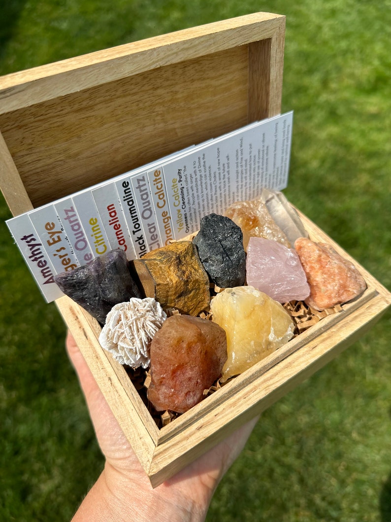 May include: A wooden box filled with various crystals and stones, including amethyst, tiger's eye, clear quartz, citrine, carnelian, black tourmaline, orange calcite, and yellow calcite. The box is open and the crystals are visible.