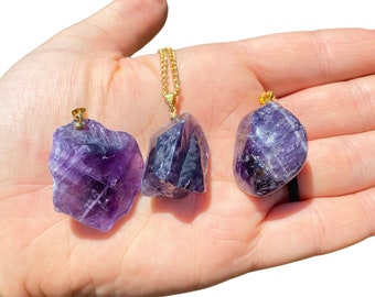 Könnte beinhalten: Drei Amethyst-Anhänger mit goldfarbenen Ösen. Ein Anhänger befindet sich an einer goldfarbenen Kette. Die Amethyststeine haben eine tiefviolette Farbe mit natürlichen Variationen in Textur und Form. Die Anhänger werden auf hellem Hintergrund präsentiert.