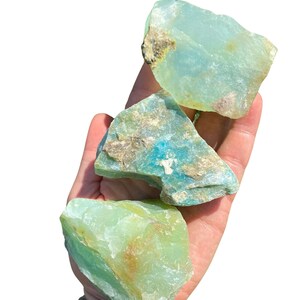 Puede incluir: Tres piedras de crisoprasa en bruto, en tonos verdes y verde azulado, se sostienen en una mano. Las piedras tienen una textura natural rugosa con zonas marrones y beige. El fondo es blanco brillante.