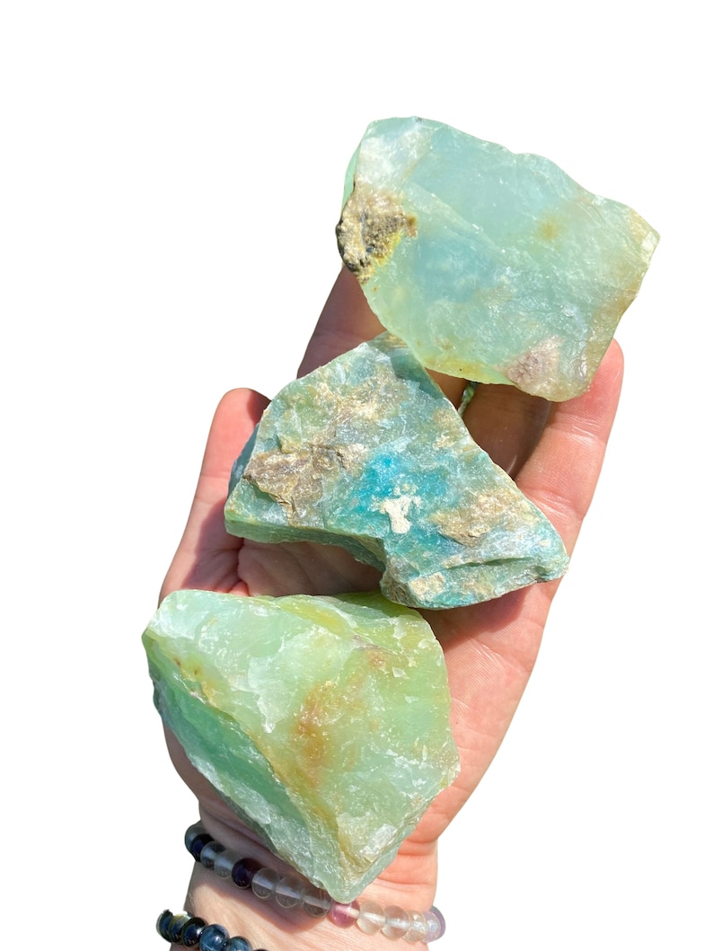 Puede incluir: Tres piedras de crisoprasa en bruto, en tonos verdes y verde azulado, se sostienen en una mano. Las piedras tienen una textura natural rugosa con zonas marrones y beige. El fondo es blanco brillante.