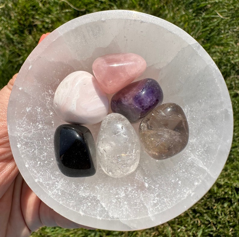 Puede incluir: Una colecci&oacute;n de seis piedras pulidas en un cuenco de selenita blanco. Las piedras son cuarzo rosa, amatista, cuarzo transparente, cuarzo ahumado, calcita rosa y obsidiana negra.