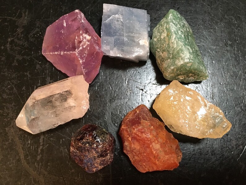 May include: A collection of seven crystals in various colours, including clear, pink, blue, green, yellow, red, and purple. The crystals are arranged in a circle on a dark surface.