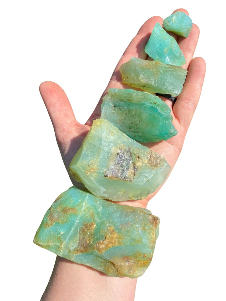 Puede incluir: Una mano sostiene varias piedras preciosas en bruto, de color verde claro. Las piedras tienen una calidad transl&uacute;cida con algunas &aacute;reas de coloraci&oacute;n marr&oacute;n y beige. Las piedras preciosas tienen forma irregular y parecen ser espec&iacute;menes naturales.