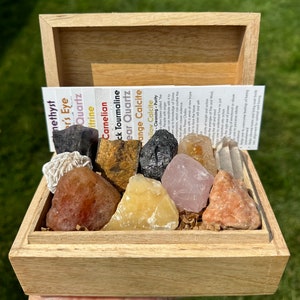 May include: A wooden box filled with various crystals and stones, including amethyst, tiger's eye, clear quartz, carnelian, black tourmaline, clear quartz, orange calcite, and yellow calcite. The box is open and the crystals are visible. The box is labeled with the names of the crystals.