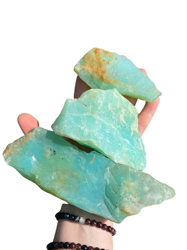 Puede incluir: Tres piezas de piedra preciosa de crisoprasa en bruto, de color verde azulado claro. Las piedras tienen una textura rugosa y natural con algunas zonas de coloraci&oacute;n marr&oacute;n y beige. Las piedras preciosas se sostienen en una mano sobre un fondo blanco.