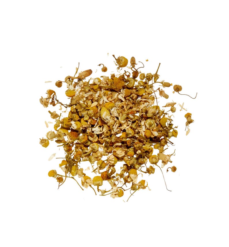 May include: A pile of dried chamomile flowers, featuring small, yellow, daisy-like blossoms and delicate stems. The dried herbs are a mix of light and dark brown, with a few white petals scattered throughout. The image is set against a clean white background.