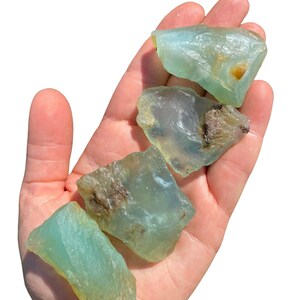 Puede incluir: Cuatro cristales en bruto, de color azul verdoso claro, se sostienen en una mano. Los cristales tienen una textura rugosa e irregular con algunas zonas marrones y amarillas. Los cristales est&aacute;n iluminados por la luz solar brillante sobre un fondo blanco.