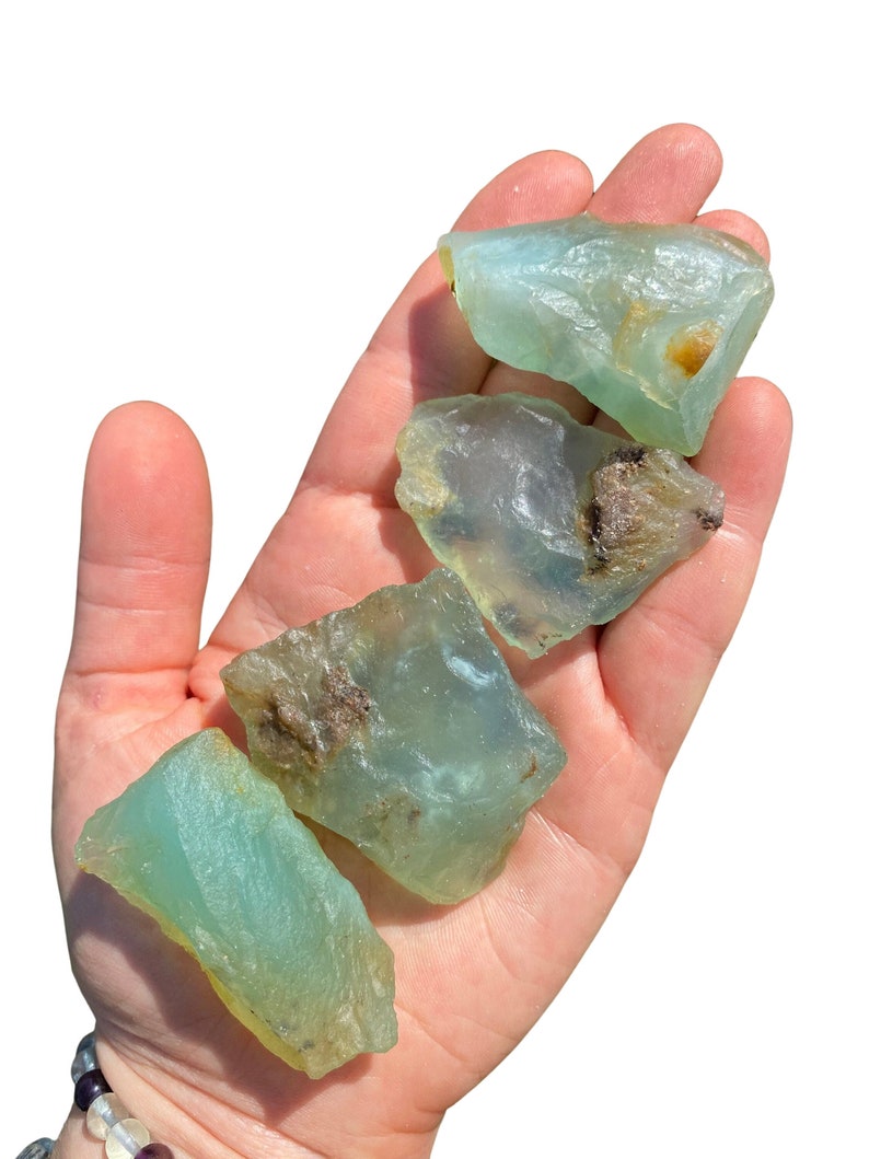 Puede incluir: Cuatro cristales en bruto, de color azul verdoso claro, se sostienen en una mano. Los cristales tienen una textura rugosa e irregular con algunas zonas marrones y amarillas. Los cristales est&aacute;n iluminados por la luz solar brillante sobre un fondo blanco.