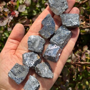 May include: A collection of ten small, gray, metallic, cubic crystals. The crystals have a rough, textured surface and are arranged in a pile on a hand.
