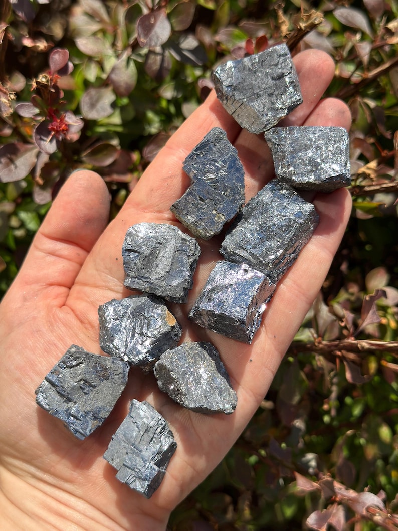 May include: A collection of ten small, gray, metallic, cubic crystals. The crystals have a rough, textured surface and are arranged in a pile on a hand.
