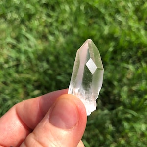 May include: A clear quartz crystal point with a natural, rough texture. The crystal is held in a hand against a green grassy background.