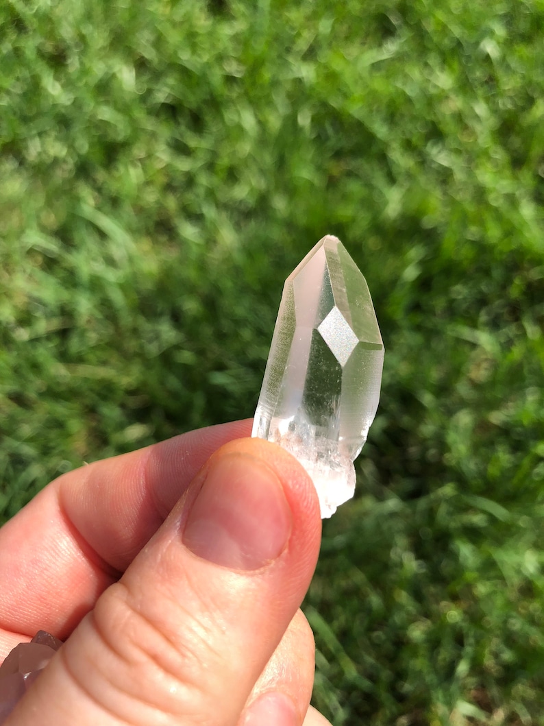 May include: A clear quartz crystal point with a natural, rough texture. The crystal is held in a hand against a green grassy background.