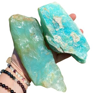 Puede incluir: Dos piedras de amazonita en bruto, de color turquesa y verde azulado, con una superficie rugosa y texturizada. Las piedras tienen &aacute;reas m&aacute;s claras y oscuras, con algunas manchas beige y blancas. Las piedras se sostienen en una mano.