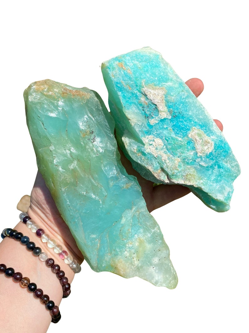 Puede incluir: Dos piedras de amazonita en bruto, de color turquesa y verde azulado, con una superficie rugosa y texturizada. Las piedras tienen &aacute;reas m&aacute;s claras y oscuras, con algunas manchas beige y blancas. Las piedras se sostienen en una mano.