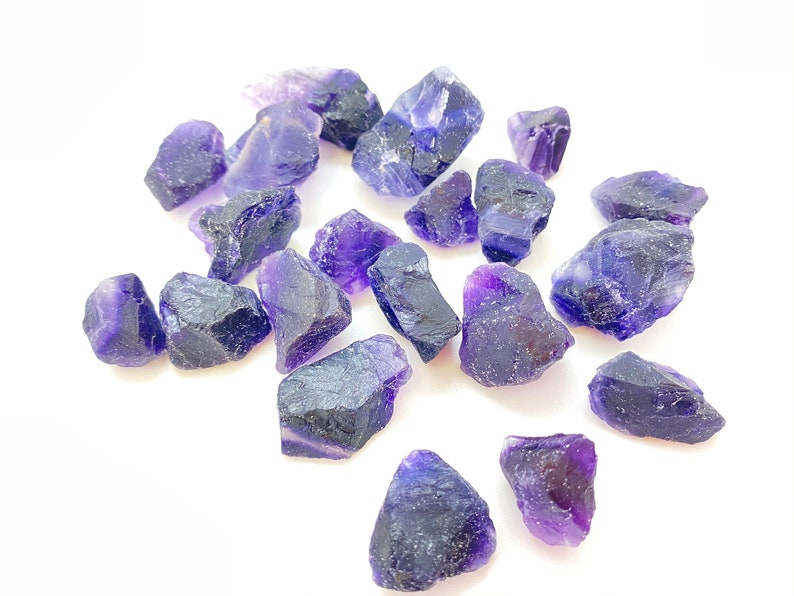 Pode incluir: Uma cole&ccedil;&atilde;o de cristais de fluorita roxa e azul, com bordas &aacute;speras e naturais. Os cristais est&atilde;o espalhados sobre um fundo branco.