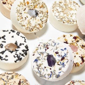 May include: Circular bath bombs with various natural decorations, including dried herbs, flower petals, and crystals. The bath bombs are white with a hole in the center, and the decorations are in shades of green, brown, pink, and blue.