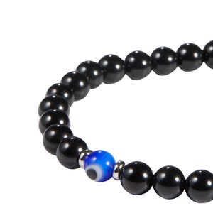 May include: Black beaded bracelet with a single blue eye bead. The bracelet features glossy, round black beads and a central blue bead with a white and black eye design. The bracelet is strung on a flexible cord.