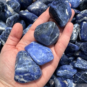 May include: A collection of smooth, polished blue and white Sodalite tumbled stones. The stones have a variety of shapes and sizes, and they are arranged in a pile. The stones are a deep blue color with white veins running through them.