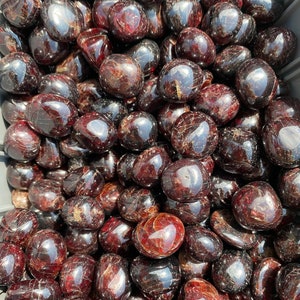 May include: A close-up image of a large pile of tumbled red garnet stones. The stones are smooth and polished, with a deep red color and a slight sparkle.
