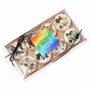 Puede incluir: Una caja rectangular que contiene ocho velas blancas redondas cubiertas con varias piedras preciosas coloridas y hierbas secas. Una etiqueta cuadrada en el centro presenta un degradado de arcoíris y el texto: NEW MOON BEGINNINGS CHAKRA MIXED PACK.