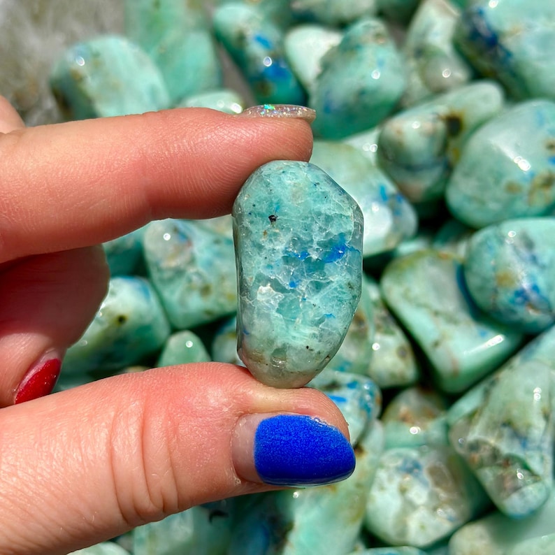 May include: A close-up of a tumbled blue green apophyllite crystal. The crystal has a light blue and green color with a white, cloudy appearance. The crystal is smooth and polished.