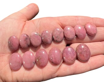 May include: A hand holding fourteen oval-shaped pink gemstones. The stones vary in shade from light to medium pink, with some showing darker veining. The gemstones are smooth and polished, reflecting light.