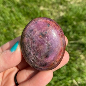 May include: A smooth, polished, pink and red ruby gemstone with a swirling pattern. The stone is oval shaped and has a glossy finish.