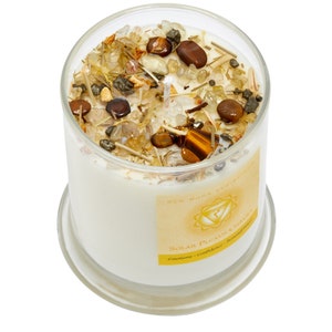 May include: A clear glass jar candle with a white wax base and a yellow label that reads "New Moon Rituals Solar Plexus Chakra Emotions - Confidence - Personal Power". The jar is filled with small stones and crystals, including tiger's eye, clear quartz, and citrine.
