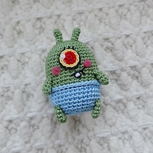 Pode incluir: Um brinquedo de pelúcia verde de crochê com um único olho vermelho grande, uma pequena boca e bochechas cor-de-rosa. O brinquedo está usando uma fralda de crochê azul. O brinquedo está sentado em um cobertor branco de crochê.
