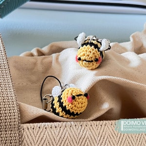 Peut inclure: Deux porte-clés en forme d'abeille en crochet avec des rayures noires et jaunes, des ailes blanches et des joues roses. Les porte-clés sont posés sur une surface en tissu beige.