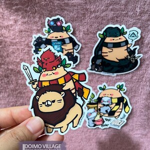 May include: Four illustrated stickers featuring a brown bear wearing a scarf and glasses, a brown bear wearing a scarf and a black hat, a brown bear wearing a scarf and riding a lion, and a brown bear wearing a scarf and holding a potion bottle. The stickers are all on a pink fuzzy surface.