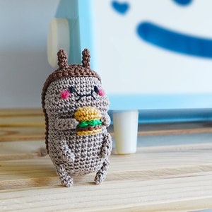 Può includere: Un orsacchiotto amigurumi di peluche marrone all'uncinetto che tiene un hamburger verde e marrone. L'orso ha un naso marrone, guance rosa e un cappello marrone all'uncinetto. L'orso è seduto su uno scaffale di legno. Il testo "DOIMOVILLAGE where handmade bearz are @etsy.doimovillage" è visibile sullo sfondo.