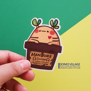 May include: A brown and tan cartoon mandrake plant with green leaves and a red heart. The plant is in a brown pot with a yellow warning label that says "Mandrake Caution".