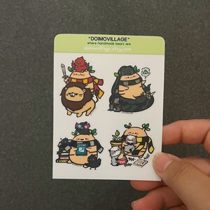 May include: Four sticker designs featuring a cartoon bear wearing a Hogwarts house scarf. The bear is wearing Gryffindor, Slytherin, Ravenclaw, and Hufflepuff scarves. The stickers are on a white background with the text "DOIMOVILLAGE" and "Where handmade bears are DOIMOVILLAGE.ETSY.COM".