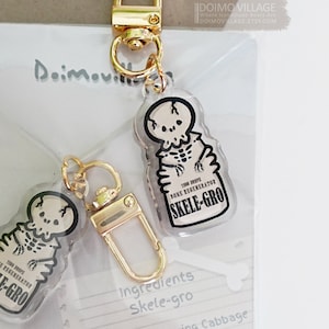 May include: A clear plastic keychain charm featuring a cartoon skeleton holding a bottle labeled "1100 Drops Bone Regenerator Skele-Gro". The charm is attached to a gold metal key ring. The image also includes a handwritten recipe for Skele-Gro, listing ingredients such as Chinese Chomping Cabbage, Puffer-fish, Scarab Beetles, and Fanged Geranium.