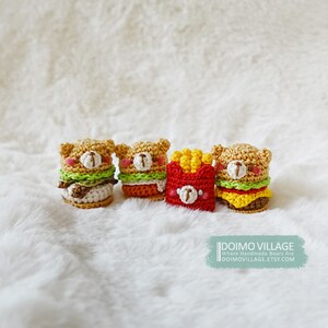 May include: Four crocheted stuffed toys in the shape of hamburgers and french fries. The hamburgers are brown, yellow, and green, and the french fries are yellow and red. The toys are arranged in a row on a white fluffy surface. The text "DOIMO VILLAGE" is visible in the bottom right corner of the image.