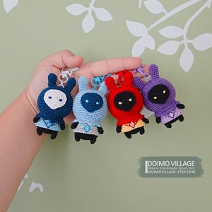 Puede incluir: Cuatro peluches de crochet con forma de conejo con cuerpos de diferentes colores: azul, azul claro, rojo y morado. Cada juguete tiene una cara negra con ojos blancos y un pequeño diseño en forma de diamante en el pecho. Los juguetes cuelgan de llaveros.