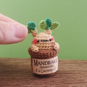 Può includere: Una pianta di mandragola all'uncinetto in un vaso marrone con un'etichetta che recita "Mandrake [Immature] CAUTION". La pianta ha foglie verdi e un corpo marrone con un viso carino.
