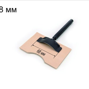 May include: A black leather working tool with a handle and a curved metal blade. The tool is placed on a piece of brown leather with a measurement of 68 mm marked on it.
